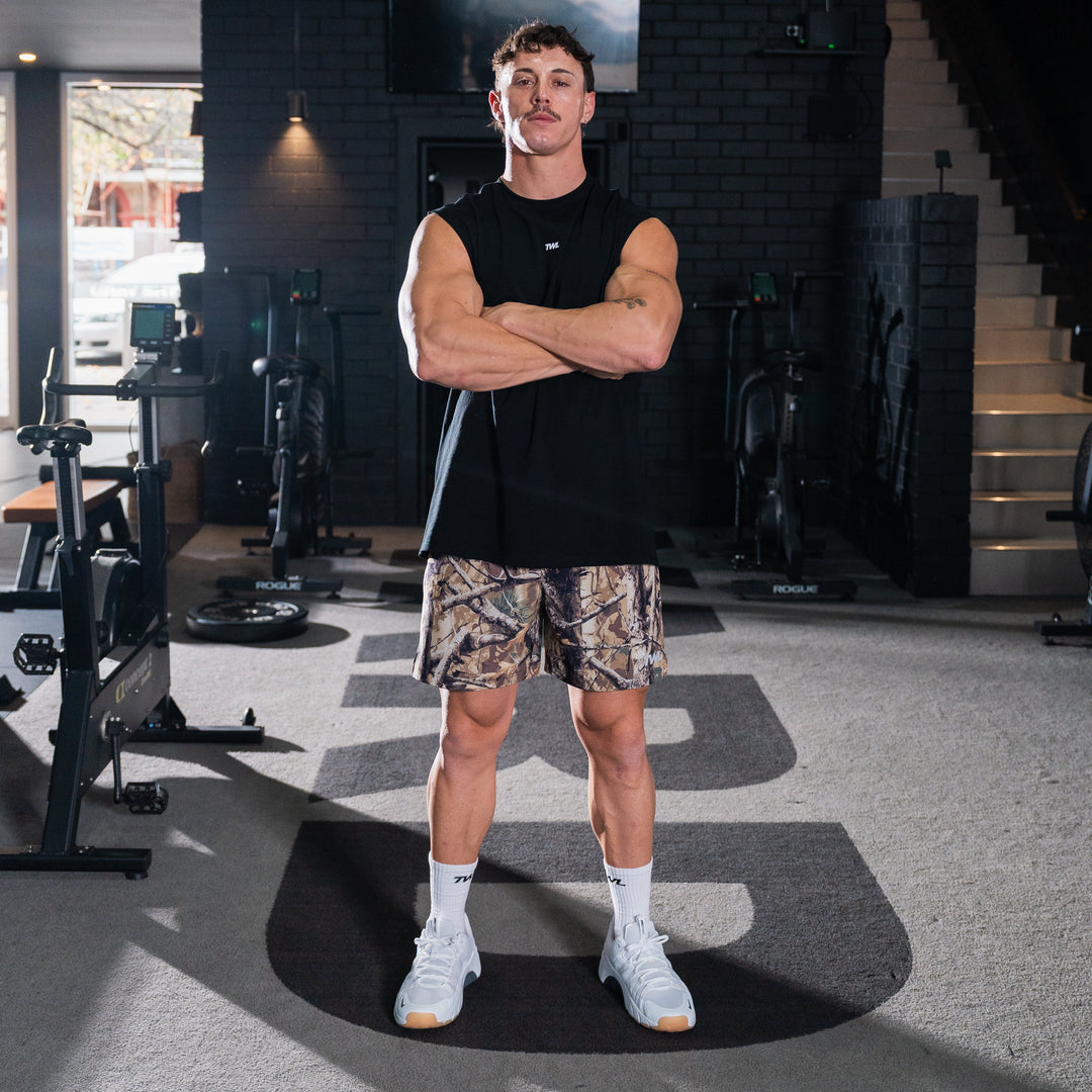 TWL Men's Vault Shorts 6 inch training shorts in tree camo, in gym with man posing wearing the shorts and a muscle tank