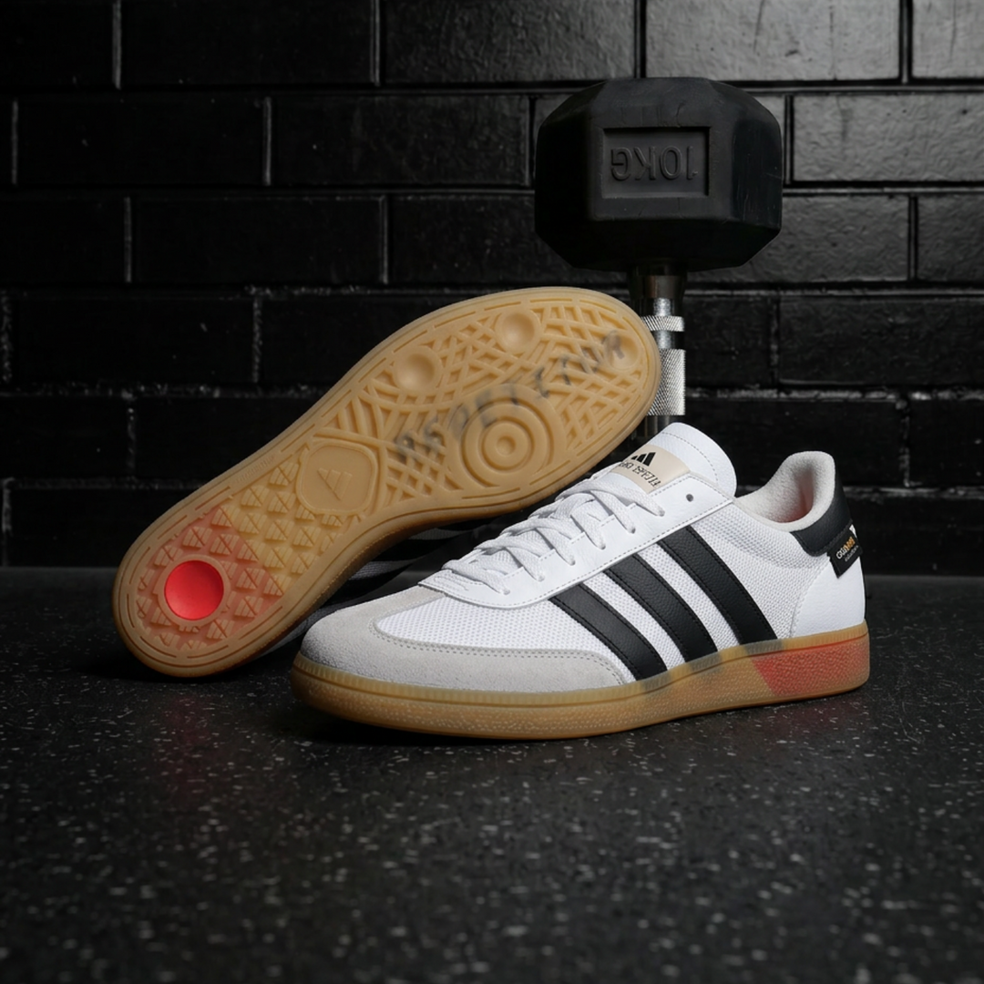 A pair of Adidas Spezial sneakers in white with black stripes, a gum sole, and a red heel accent, displayed on a dark textured floor with a black brick wall and a dumbbell in the background.