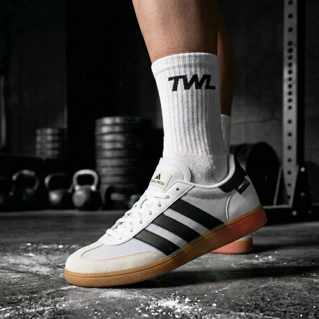 A person wearing a white Adidas Spezial sneaker with black three-stripes and a gum sole, paired with white crew socks featuring the black TWL logo, in a gym setting.