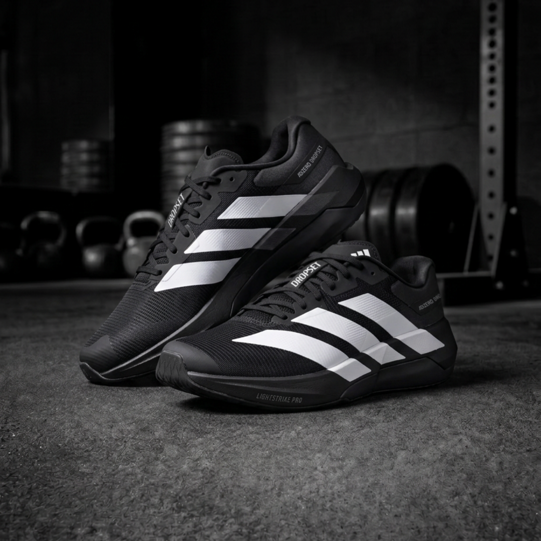 A pair of black Adidas Dropset Pro shoes with white three-stripes, displayed on a dark gym floor with weights in the background.