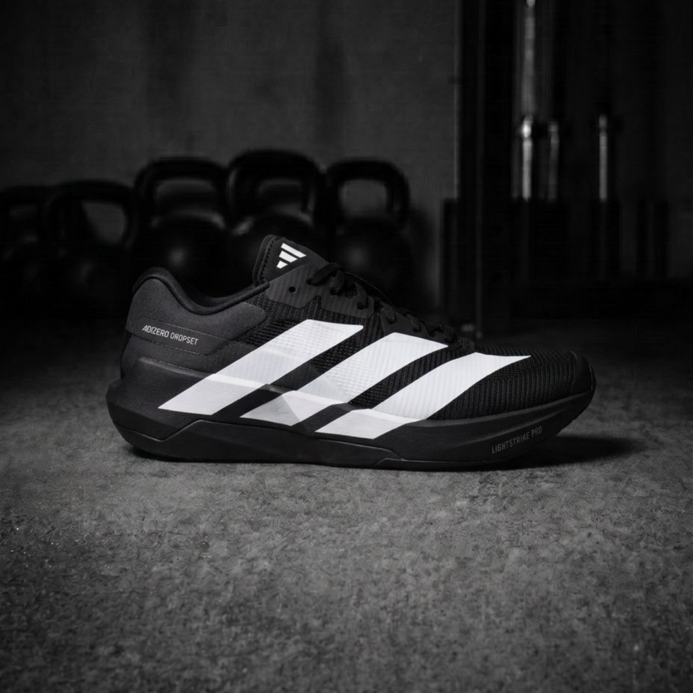 A black Adidas Ultraboost running shoe with three white stripes, positioned on a dark concrete floor with kettlebells blurred in the background.