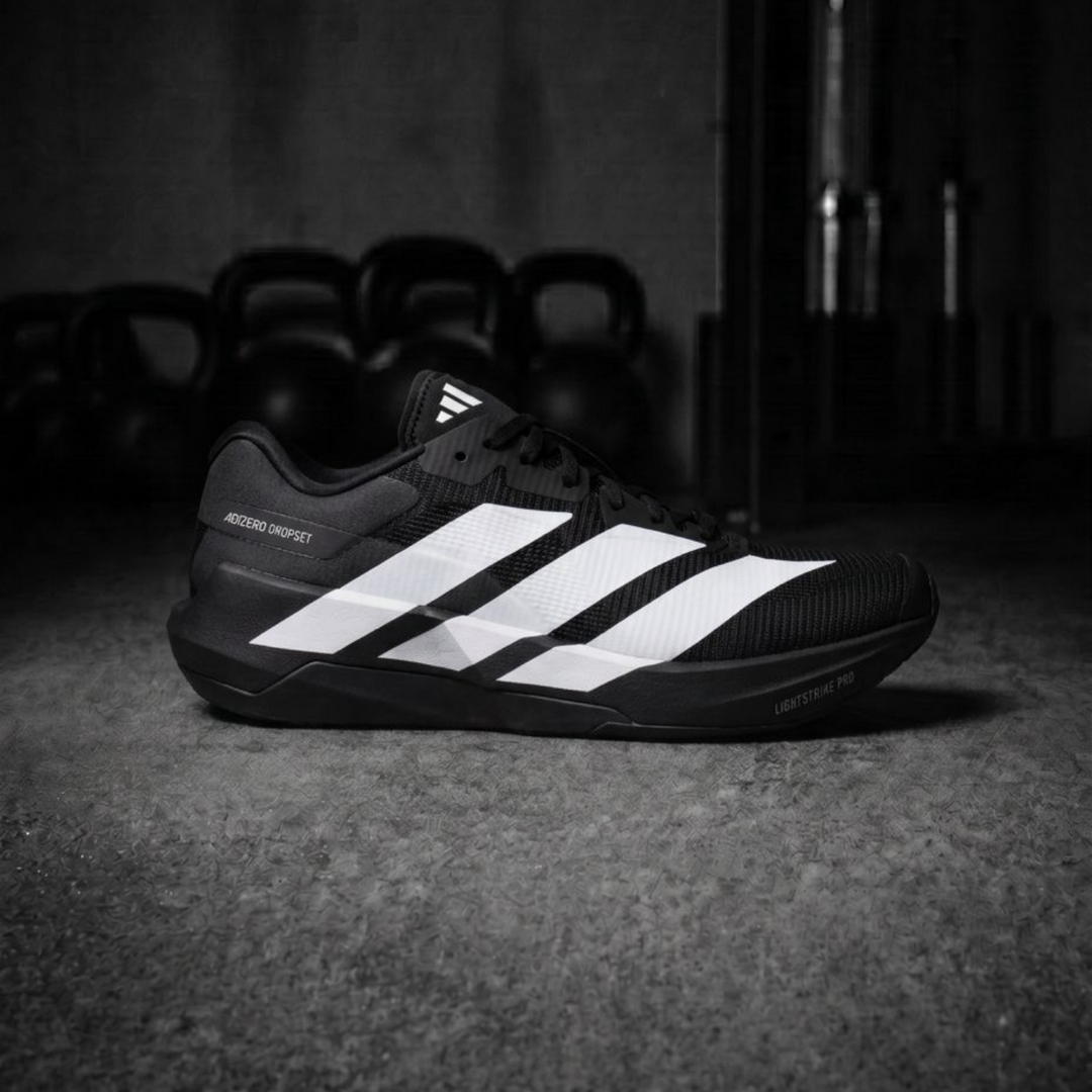 A black Adidas Ultraboost running shoe with three white stripes, positioned on a dark concrete floor with kettlebells blurred in the background.