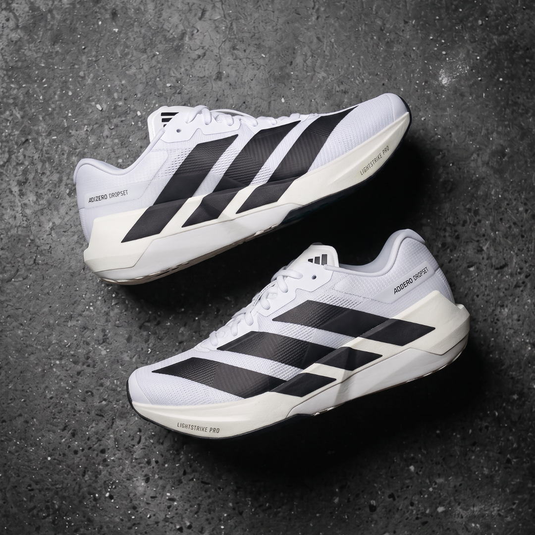 A pair of Adidas Dropset Pro running shoes in white and black, arranged diagonally on a dark textured concrete surface.