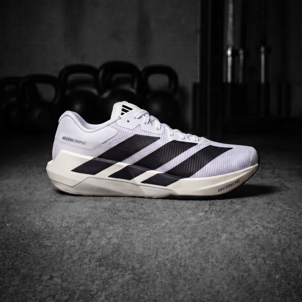 A single Adidas Dropset Pro running shoe in white, black, and off-white, positioned in a gym setting with kettlebells blurred in the background.