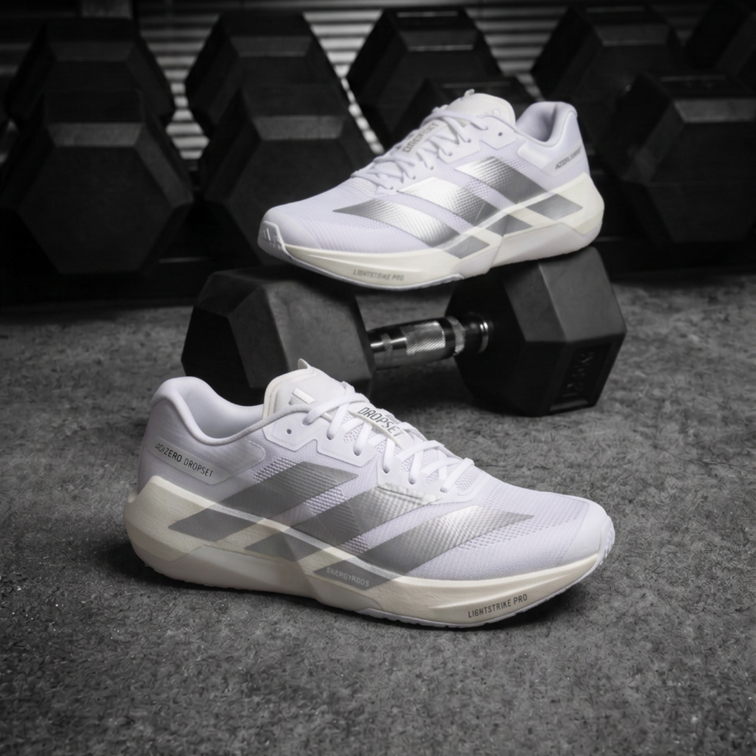 A pair of white and silver Adidas Dropset Pro athletic sneakers displayed on and around black hex dumbbells on a dark gray textured floor, with a blurred gym background.