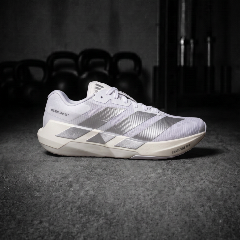 A single Adidas Dropset Pro running shoe in white and silver, positioned on a dark textured floor with kettlebells blurred in the background.