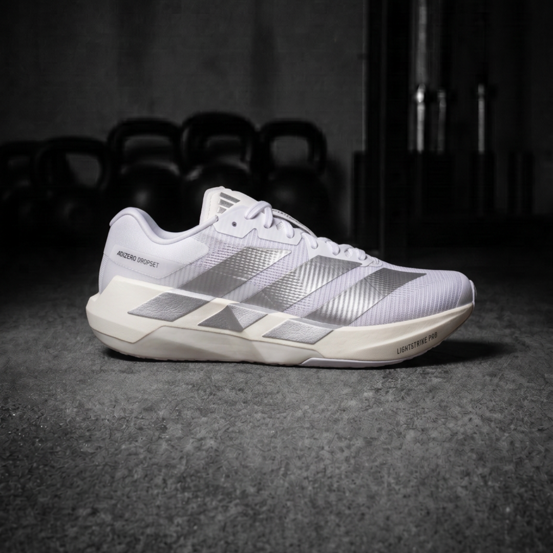 A single Adidas Dropset Pro running shoe in white and silver, positioned on a dark textured floor with kettlebells blurred in the background.
