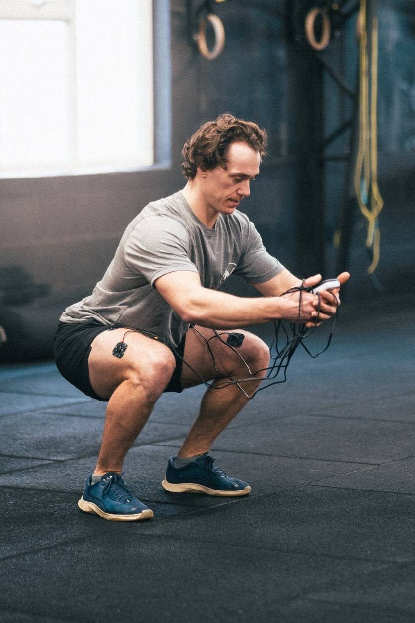A man squatting in a gym, using the Compex SP 2.0 EMS muscle stimulator for a quad muscle strengthening session