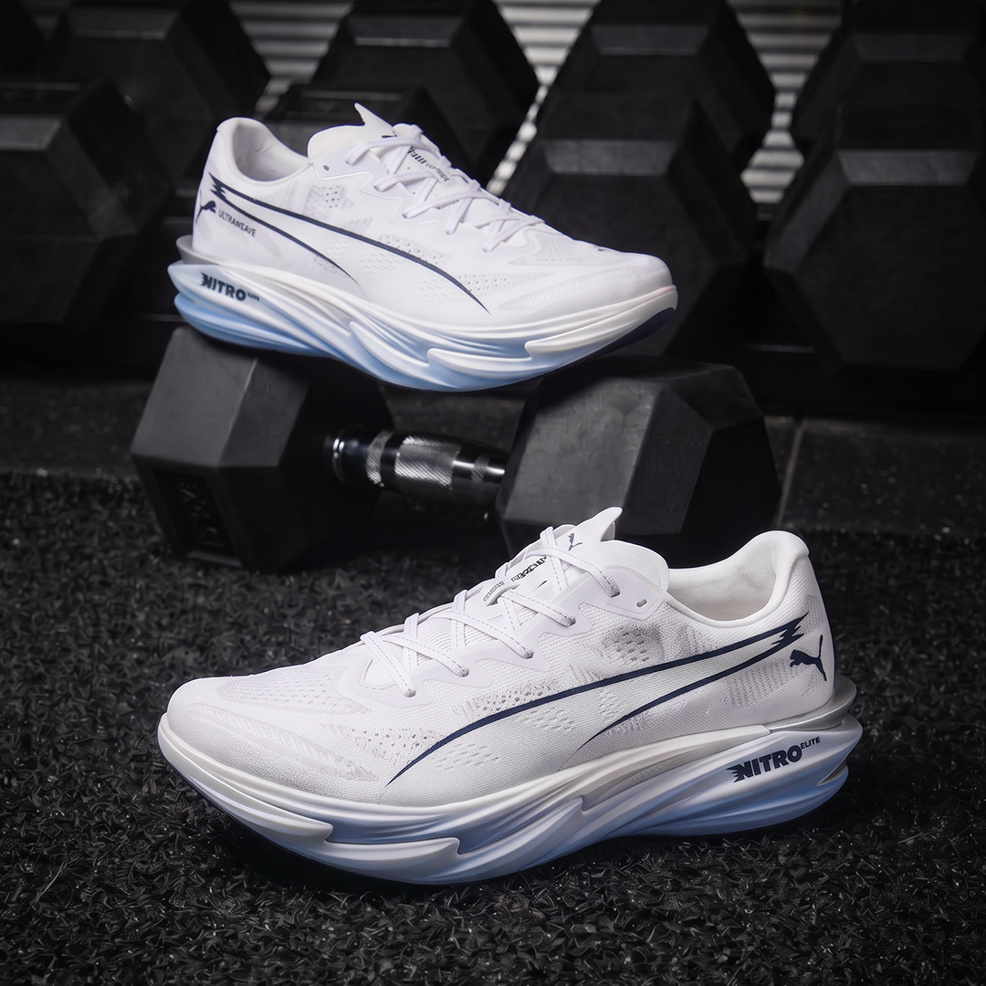 A pair of white Puma Nitro running shoes with navy blue accents and translucent blue midsoles, displayed on a dark textured floor with black dumbbells in the background.
