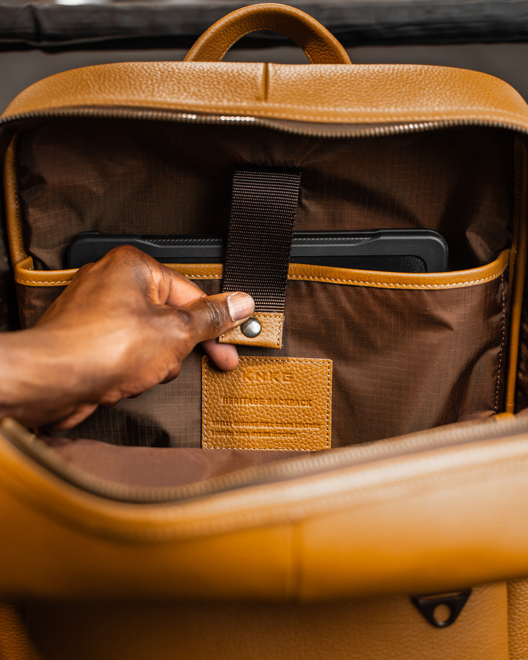 Man reaching for laptop inside of sleeve of tan leather KNKG HERITAGE Backpack