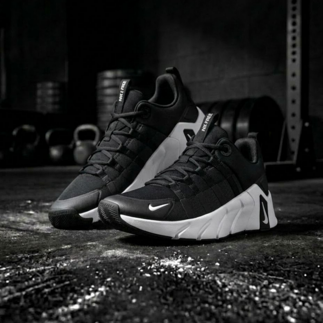 A pair of Nike sneakers in black and white, positioned on a dark, gritty gym floor with weight equipment blurred in the background.