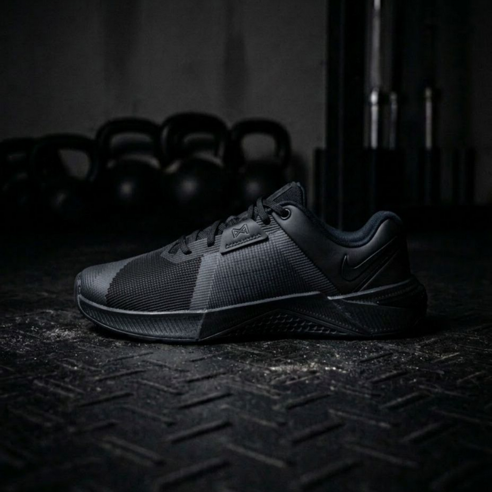 A black Nike athletic shoe positioned on a dark textured metal floor, with blurred kettlebells in the background.