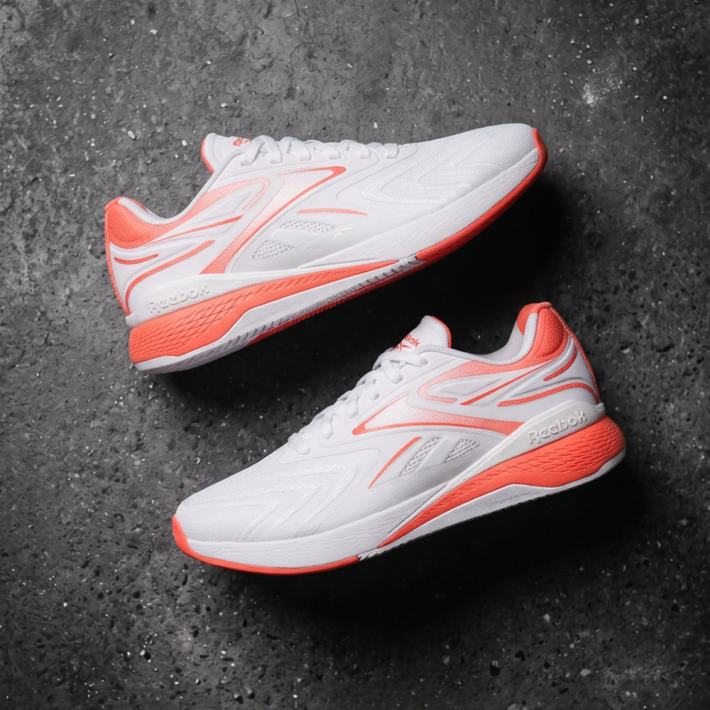 A pair of white athletic Reebok sneakers with vibrant orange accents, displayed diagonally on a dark textured concrete surface. 