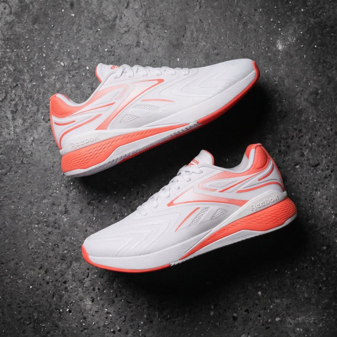 A pair of white athletic Reebok sneakers with vibrant orange accents, displayed diagonally on a dark textured concrete surface. 