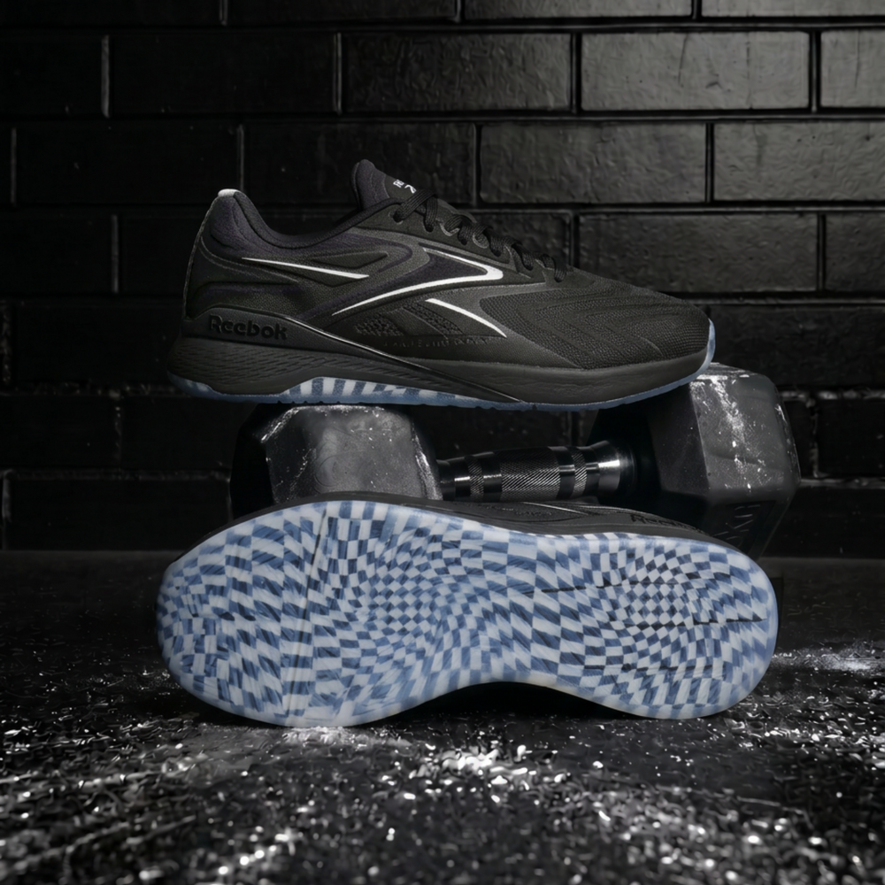 A Reebok athletic shoe in dark gray and black, displayed with one shoe standing upright and the other lying on its side to show the blue and white checkered outsole, set against a dark brick wall background.