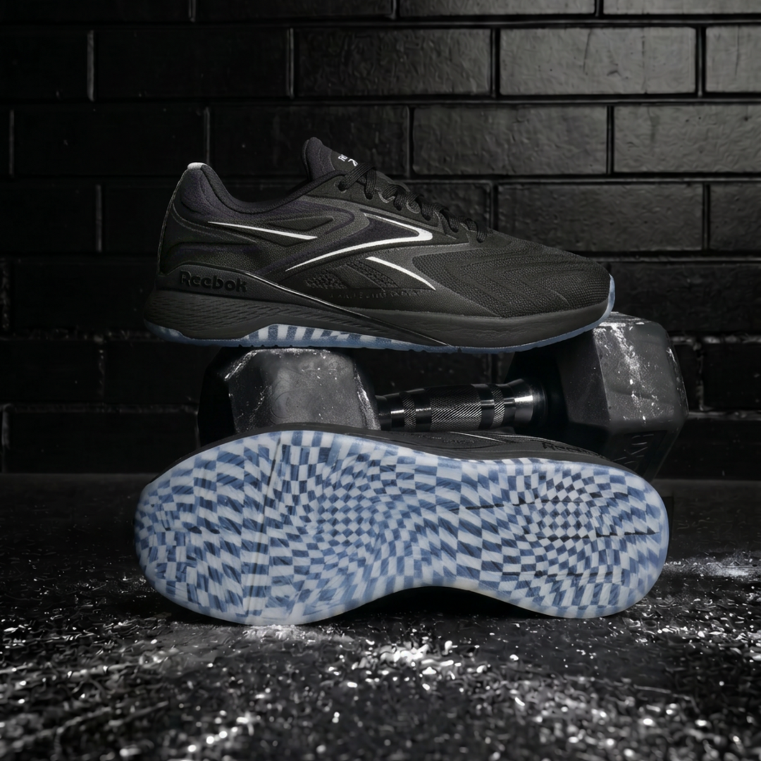A Reebok athletic shoe in dark gray and black, displayed with one shoe standing upright and the other lying on its side to show the blue and white checkered outsole, set against a dark brick wall background.