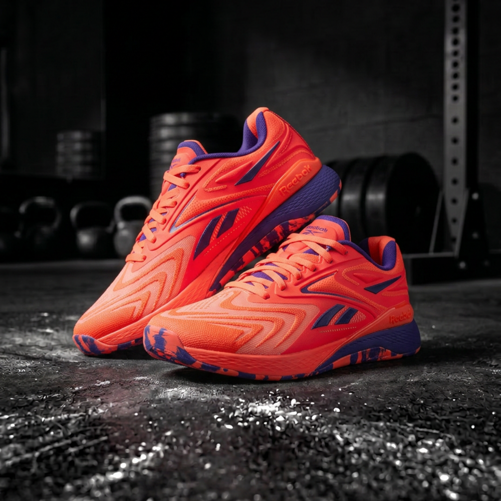 A pair of Reebok running shoes in a vibrant neon orange and purple colorway, displayed on a dark, gritty gym floor with weight plates blurred in the background.