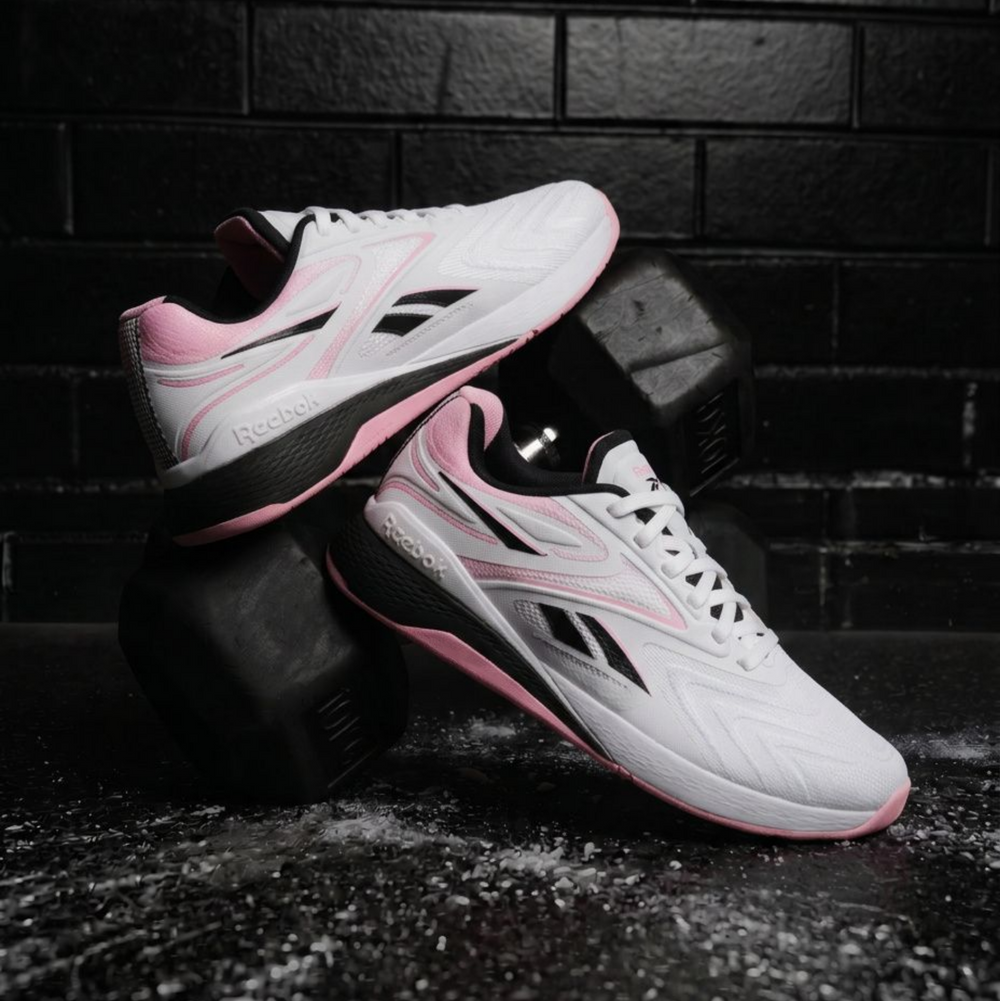 A pair of Reebok sneakers in white, black, and pink, styled on top of a black dumbbell against a dark brick wall background.