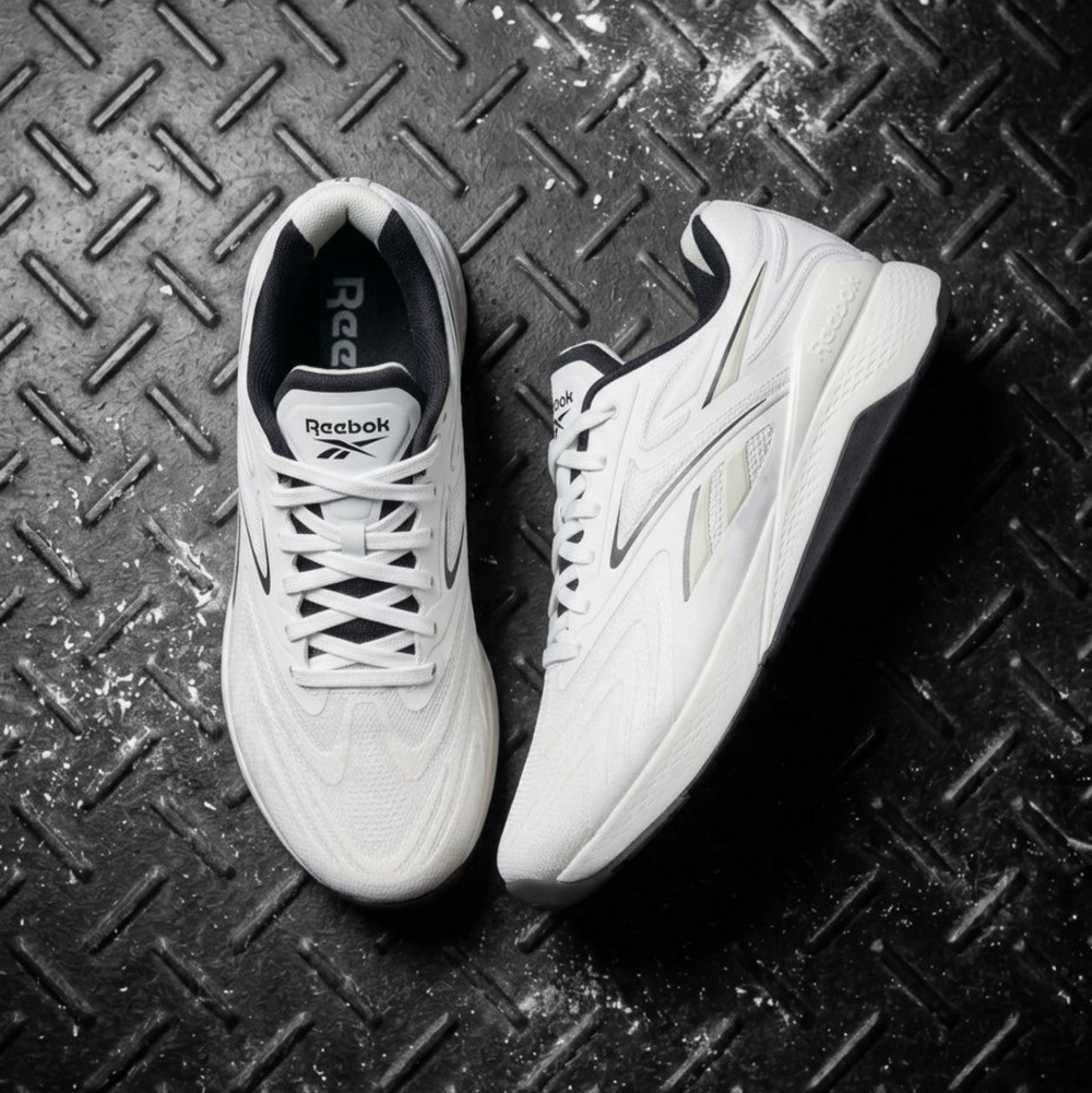 A pair of Reebok athletic sneakers in white with black and gray accents, displayed on a textured black diamond plate background.